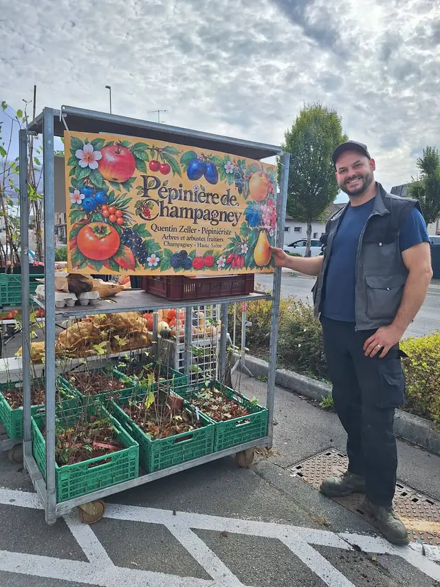 Le pépiniériste devant le stand de la pépinière : une grande enseigne illustrée de fruits et, dessous, des caisses de jeunes plants.